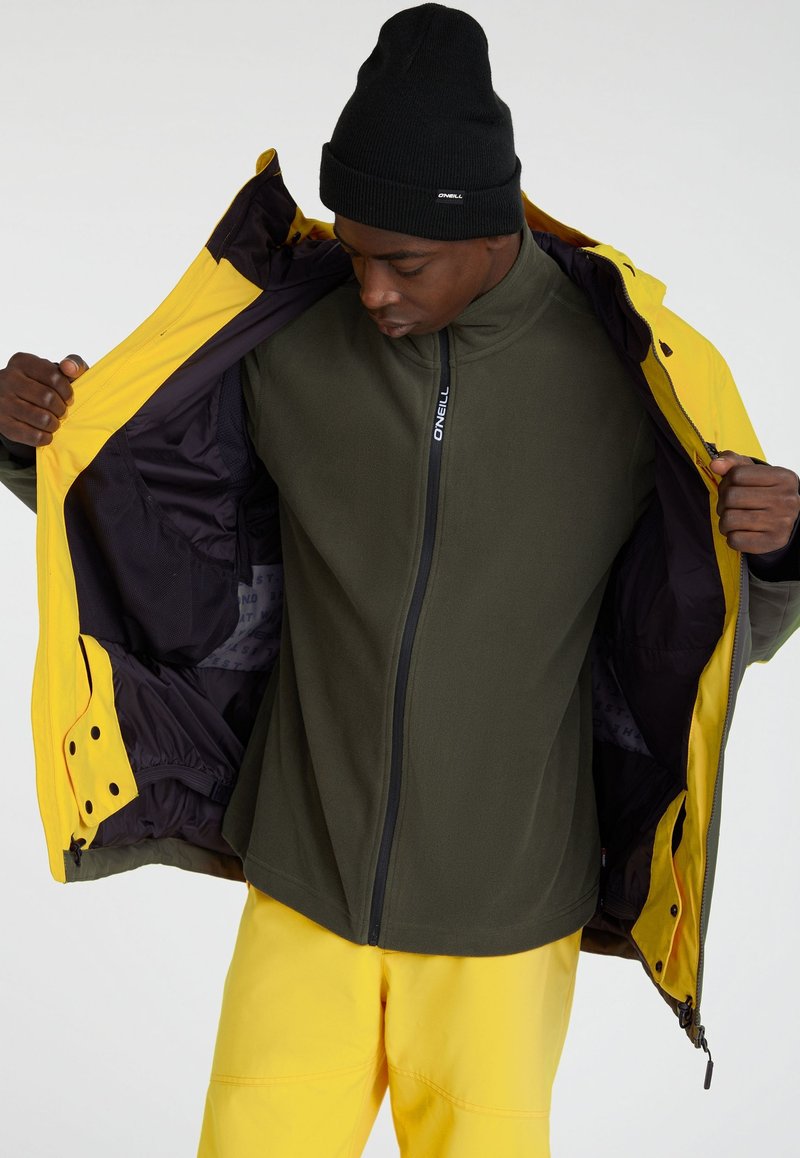 Żółta kurtka z czarnymi akcentami, rozpięta, aby ujawnić ciemnozieloną polarową podszewkę. Model nosi czarną czapkę i żółte spodnie.