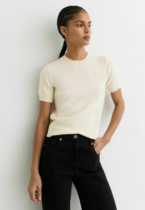 Jeune femme aux cheveux tressés portant un pull crème à manches courtes et un pantalon noir taille haute, debout contre un mur blanc.