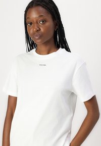 Hvit bomull T-skjorte med korte ermer og rund hals. Har en liten svart "Calvin Klein" logo på brystet. Glatt tekstur.
