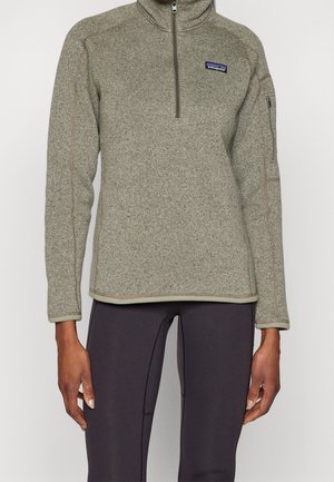 Heathergrijze fleece met een kwart rits en een ritssluiting op de mouw, gedragen over zwarte leggings bij een staand persoon.