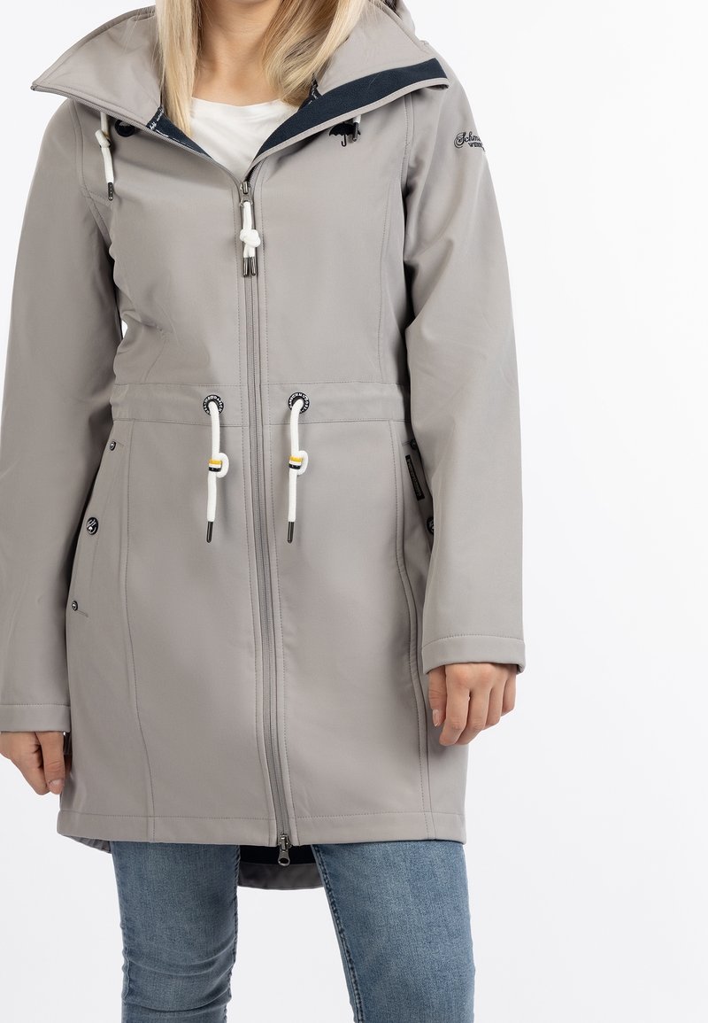 Femme portant un manteau à capuche gris clair avec une fermeture éclair avant et des cordons blancs, assorti à un jean bleu.