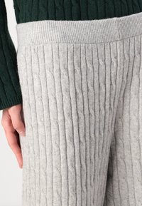 Pantaloni in maglia grigio chiaro con una texture a coste, dotati di una comoda vita elastica ampia e di un motivo a treccia verticale, abbinati a un pullover verde scuro.