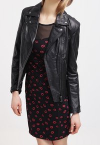 Veste de biker en cuir noir avec quincaillerie en argent, portée sur une robe noire ornée d'impressions de lèvres rouges. Texture lisse, design ajusté et fermeture éclair.