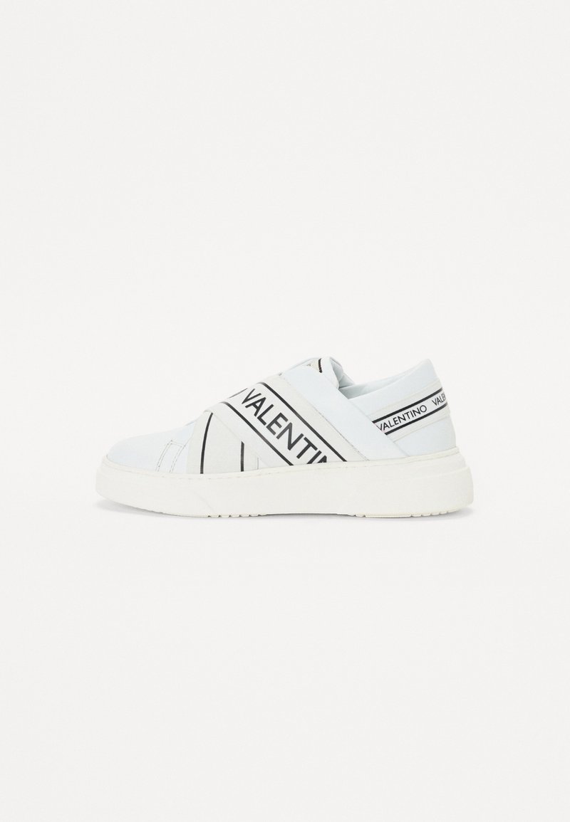 Sapatilhas brancas de slip-on com uma marcação preta, apresentando riscas diagonais e uma sola texturizada. Material liso com um design minimalista.