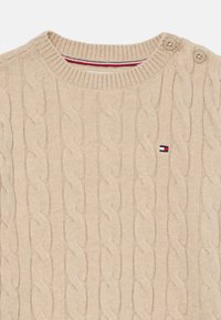 Pull en tricot beige avec un motif en câble, un col rond côtelé et deux accents de boutons sur l'épaule. Petit logo près de l'ourlet.