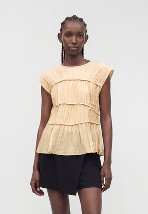 Femme portant un haut plissé beige sans manches et une jupe noire asymétrique, debout devant un fond clair uni.