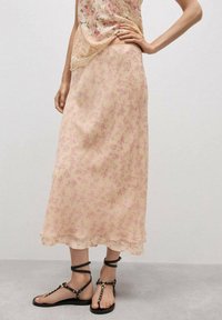 Femeie îmbrăcată cu o fustă midi bej semi-transparentă cu modele florale roz și sandale plate negre cu bretele la gleznă, stând pe un podea gri.