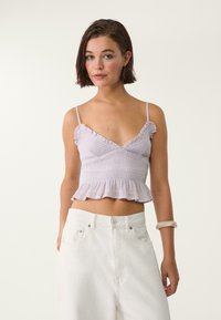 Ljubičasti crop top s volanima na naramenicama i smočnom teksturom, sa proširenim rubom. Uparen s bijelim hlačama visokog struka.