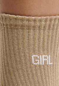 Calcetines dorados brillantes hasta la rodilla con textura de canalé; presentan el texto bordado en blanco "GIRL" en el lado exterior.