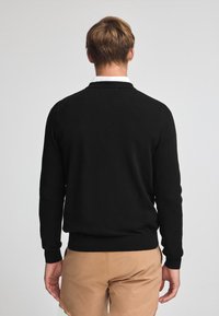 Suéter negro de material suave, con cuello redondo, mangas largas, puños y cintura acanalados. Lleva puesto sobre pantalones cortos beige.