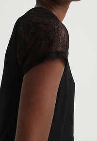 Black top featuring short sleeves with intricate black lace detailing. Smooth fabric with a relaxed fit and rounded neckline.