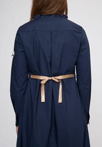 Tengerészkék, hosszú ujjú ruha fodros gallérral és bézs szatén derékövvel megkötve, sima textúrával és szabott fazonnal.