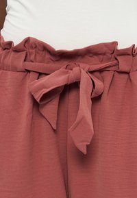 Παντελόνι ψηλόμεσο σε σκουριά απόχρωση με υφασμάτινη ζώνη δεμένη σε φιόγκο, φορεμένο με ένα λευκό τοπ μέσα στη μέση.