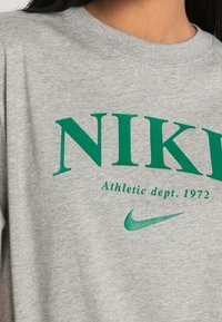 Camiseta de algodón gris con el texto verde en negrita "NIKE", "Athletic dept. 1972" en un tamaño de fuente más pequeño, y el diseño del logo de la marca Nike.