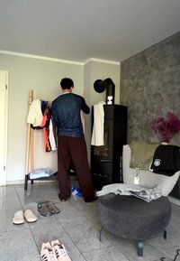 Personne en vêtements décontractés arrange des vêtements sur un porte-vêtements en bois à côté d'un poêle noir dans un espace de vie moderne avec des chaussures au sol.