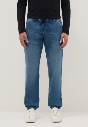 Mann trägt blaue Jogginghosen aus Denim mit elastischen Bündchen und Kordelzug am Bund, kombiniert mit weißen Turnschuhen und einem schwarzen Langarmshirt.