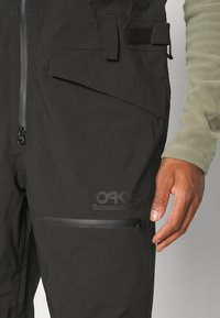 Black thermal outerwear with zippered pockets, featuring a smooth texture and a logo. Adjustable straps and a close-fit design are visible.