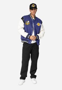 Μπουφάν varsity σε μωβ και άσπρο με κεντημένα λογότυπα, μαύρες δερμάτινες λεπτομέρειες και ριγέ μανσέτες, σε συνδυασμό με μαύρο παντελόνι και λευκά αθλητικά παπούτσια.