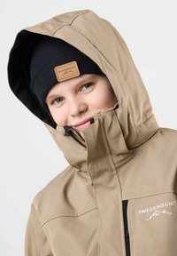 Beige Outdoor-Jacke mit schwarzem Futter und Logo. Verfügt über eine Kapuze, einen durchgehenden Reißverschluss und zwei Reißverschlusstaschen. Getragen mit einer schwarzen Strickmütze.