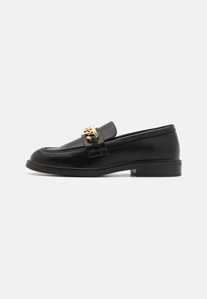 Mocassin en cuir noir avec petit talon et détail chaîne dorée sur le dessus, montré de profil sur un fond clair uni.