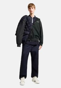 Giacca a vento nera appoggiata su una felpa con zip verde scuro, abbinata a pantaloni blu navy e sneakers bianche, in piedi di fronte a uno sfondo chiaro.