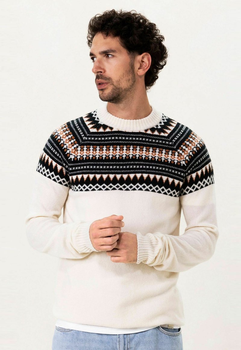 Pull en tricot avec une base crème, présentant un empiècement géométrique noir, orange et blanc. Col et poignets côtelés.