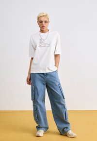 T-shirt en coton blanc avec une illustration d'une paire de baskets et de fleurs, associé à un pantalon cargo baggy bleu clair et des baskets beiges.