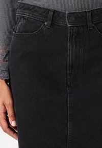 Jupe en denim noir avec une fermeture à boutons sur le devant, deux poches latérales et une silhouette droite, présentant des détails de texture et de couture subtils.