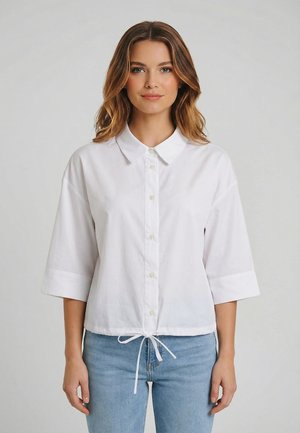 Giovane donna con capelli castani ondulati che indossa una camicia bianca con bottoni e maniche a tre quarti e jeans azzurri, in piedi davanti a uno sfondo semplice.