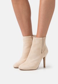 Bottines en daim beige avec un bout pointu et un talon haut élancé, dotées d'une fermeture éclair latérale et d'une texture lisse.
