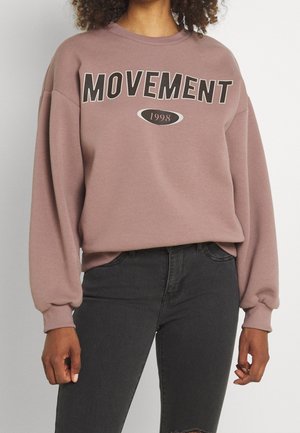 Roze sweatshirt met "MOVEMENT" in het zwart gedrukt, voorzien van een geribbelde ronde hals en lange mouwen, gecombineerd met donkere jeans.