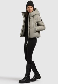 Kvinde iført grå puffjakke, sort hue, sorte leggings og knæhøje sorte støvler, stående mod en ensfarvet lys baggrund.