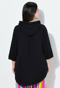 Felpa nera con cappuccio realizzata in tessuto testurizzato, con maniche a tre quarti e orlo curvo. Il modello indossa pantaloni a righe multicolori.