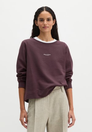Femme aux longs cheveux foncés portant un sweat-shirt violet Marc O'Polo par-dessus une chemise blanche et un pantalon beige, debout devant un fond uni.