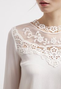 Blusa bianca con un collo in pizzo trasparente caratterizzato da ricami floreali, tessuto strutturato e maniche lunghe, che offre un aspetto delicato ed elegante.