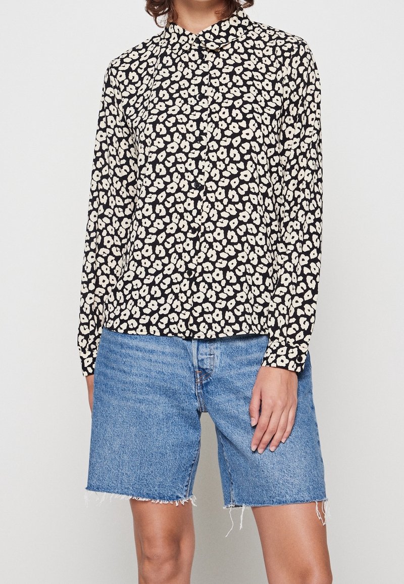 Blusa nera con motivo floreale bianco, colletto, maniche lunghe; abbinata a pantaloncini di jeans blu, orlo sfrangiato, fit rilassato.