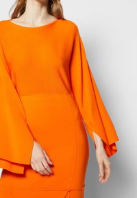 Orange långärmad klänning med en lös, fladdermusdesign. Slät textur, kroppsnära silhuett och subtila sömdetaljer vid midjan.