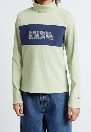 Personne portant un col roulé côtelé vert clair avec un panneau bleu marine et un texte blanc à motif "NIKE", associé à un jean bleu.