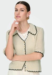 Pletený kardigan v krémové barvě s černým lemem, krátké rukávy, kulatý límec a zapínání na knoflík, který má celoplošný strukturovaný vzor.