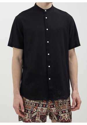 Homme portant une chemise noire à manches courtes avec col mandarin boutonnée jusqu'en haut et un short à motifs géométriques dans des tons chauds.
