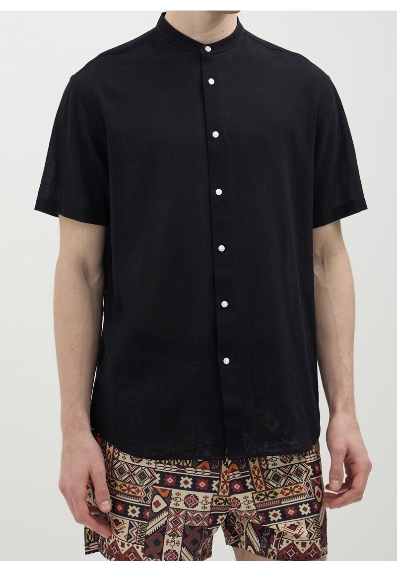 Homme portant une chemise noire à manches courtes avec col mandarin boutonnée jusqu'en haut et un short à motifs géométriques dans des tons chauds.