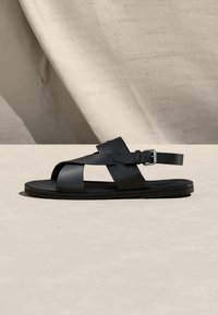 Μαύρο δερμάτινο σανδάλι με διασταυρούμενους ιμάντες και ασημένια αγκράφα σε επίπεδη σόλα, εκτεθειμένο σε μπεζ επιφάνεια με υφασμάτινο φόντο.