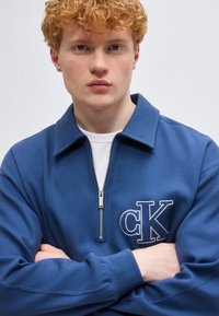 Νεαρός με σγουρά κόκκινα μαλλιά που φοράει μπλε φούτερ με φερμουάρ στη μέση και λογότυπο "cK", με σταυρωμένα τα χέρια, κοιτάζοντας μπροστά σε απλό φόντο.
