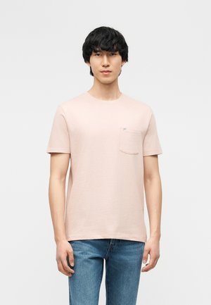 Młody mężczyzna stoi, ubrany w jasnoróżową koszulkę z krótkim rękawem z kieszenią na piersi oraz niebieskie dżinsy, na jednolitym białym tle.