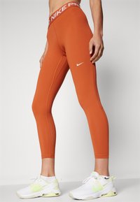 Pomarańczowe legginsy o dopasowanym kroju, z elastycznego materiału i szerokim pasem w talii z napisem "NIKE PRO" wydrukowanym na biało. Małe białe logo po boku.
