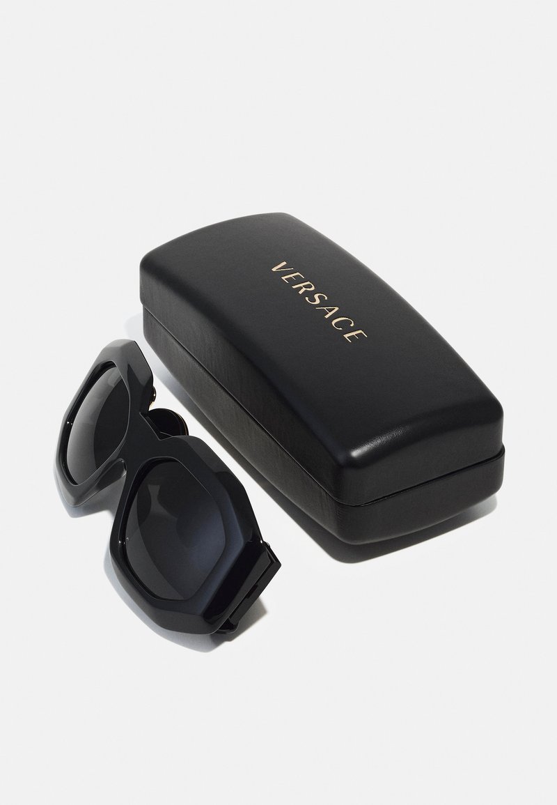 Occhiali da sole neri oversize con lenti scure, forma ottagonale e montatura nera chunky, posizionati accanto a una custodia in pelle nera impressa con "VERSACE" in oro.