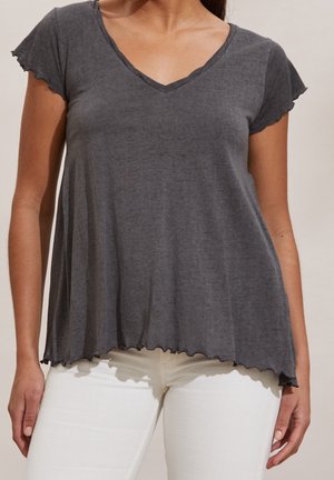 Femme portant un haut gris foncé à manches courtes avec un col en V et des bords festonnés, ainsi qu'un pantalon blanc, debout devant un fond uni.