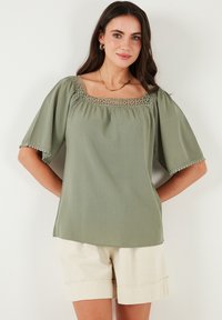 Olive green blouse with a square neckline, lace trim, and short puff sleeves. Lightweight fabric, paired with beige shorts.