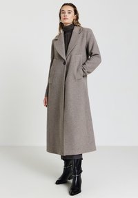 Donna in piedi che indossa un lungo cappotto di lana taupe sopra un maglione dolcevita scuro, pantaloni scuri e stivali neri con tacco.
