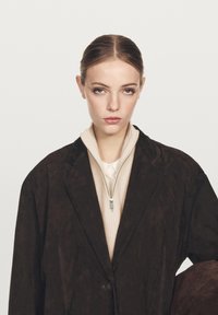 Brown suede oversized coat with a notch lapel, layered over a beige zip-up sweater. The collar is high, and accentuated by a zip closure.